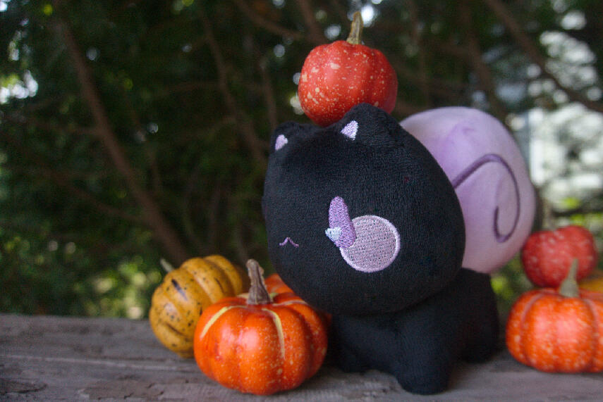 A black plush snail-cat with a purple shell surrounded by small faux pumpkins